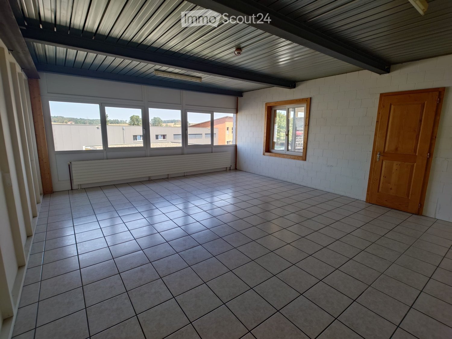 Empty room with tiled floor, white brick walls, large windows, and a wooden door.