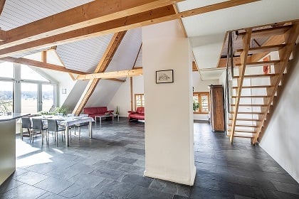 Open-plan living and dining area with exposed wooden beams, modern furniture including a red sofa, dining table with chairs, and a small stool.