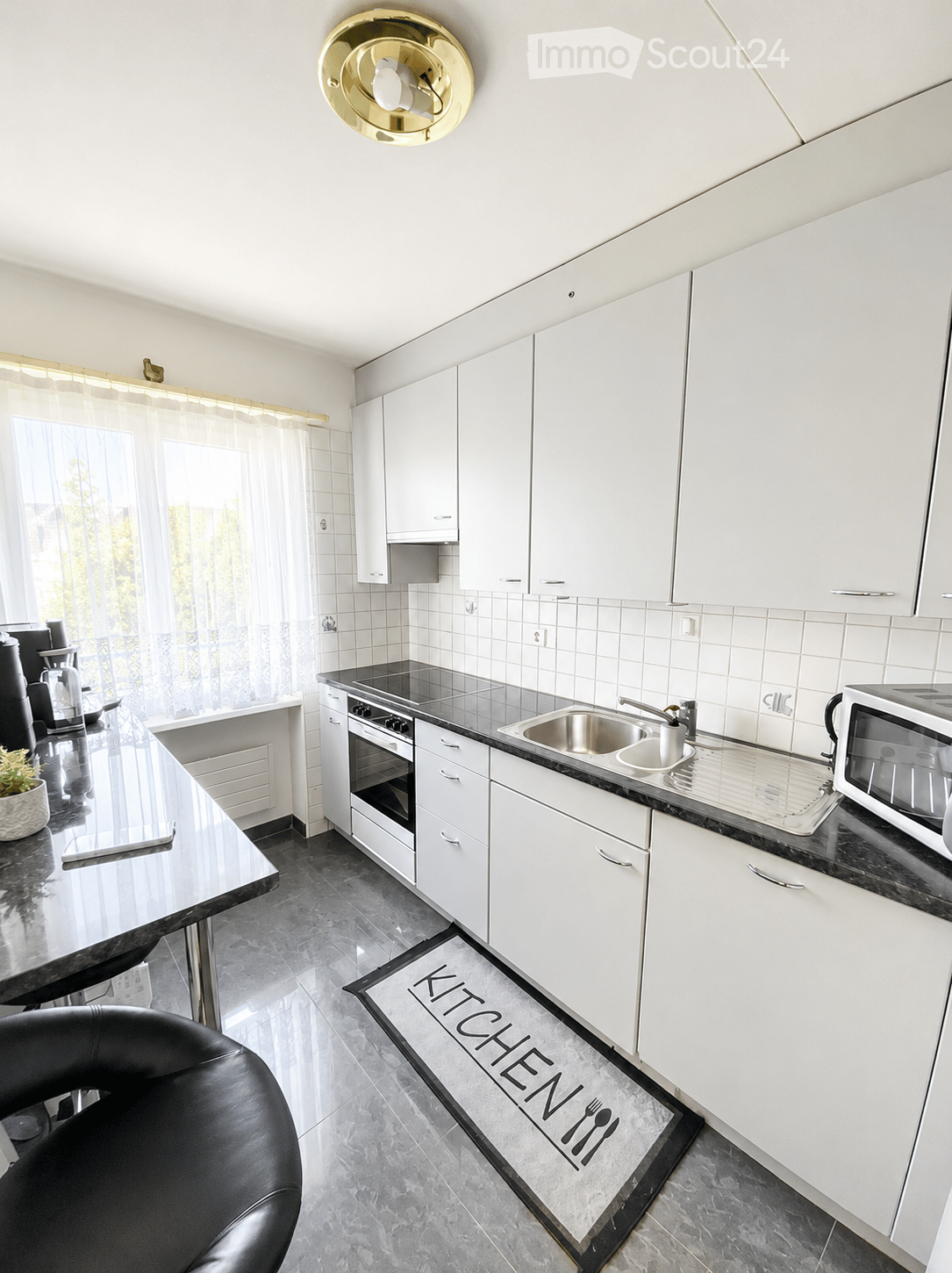 Modern kitchen with white cabinets, black countertops, stainless steel sink, oven, microwave, and a small table with a chair. A decorative rug with the word "KITCHEN" is on the floor.