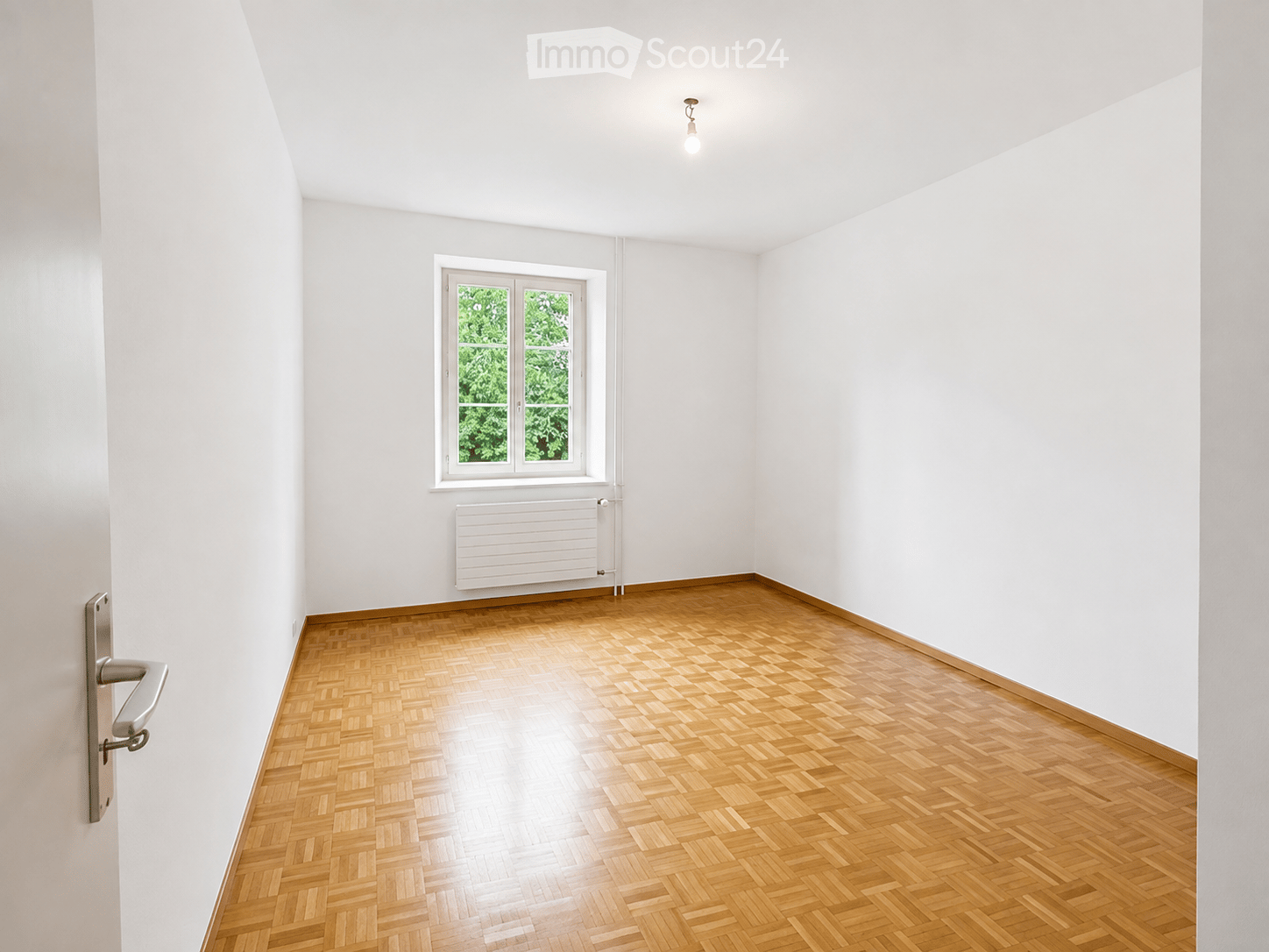 Empty room with wooden parquet flooring, white walls, a single window with white frame, radiator, and a door handle visible on the left side.