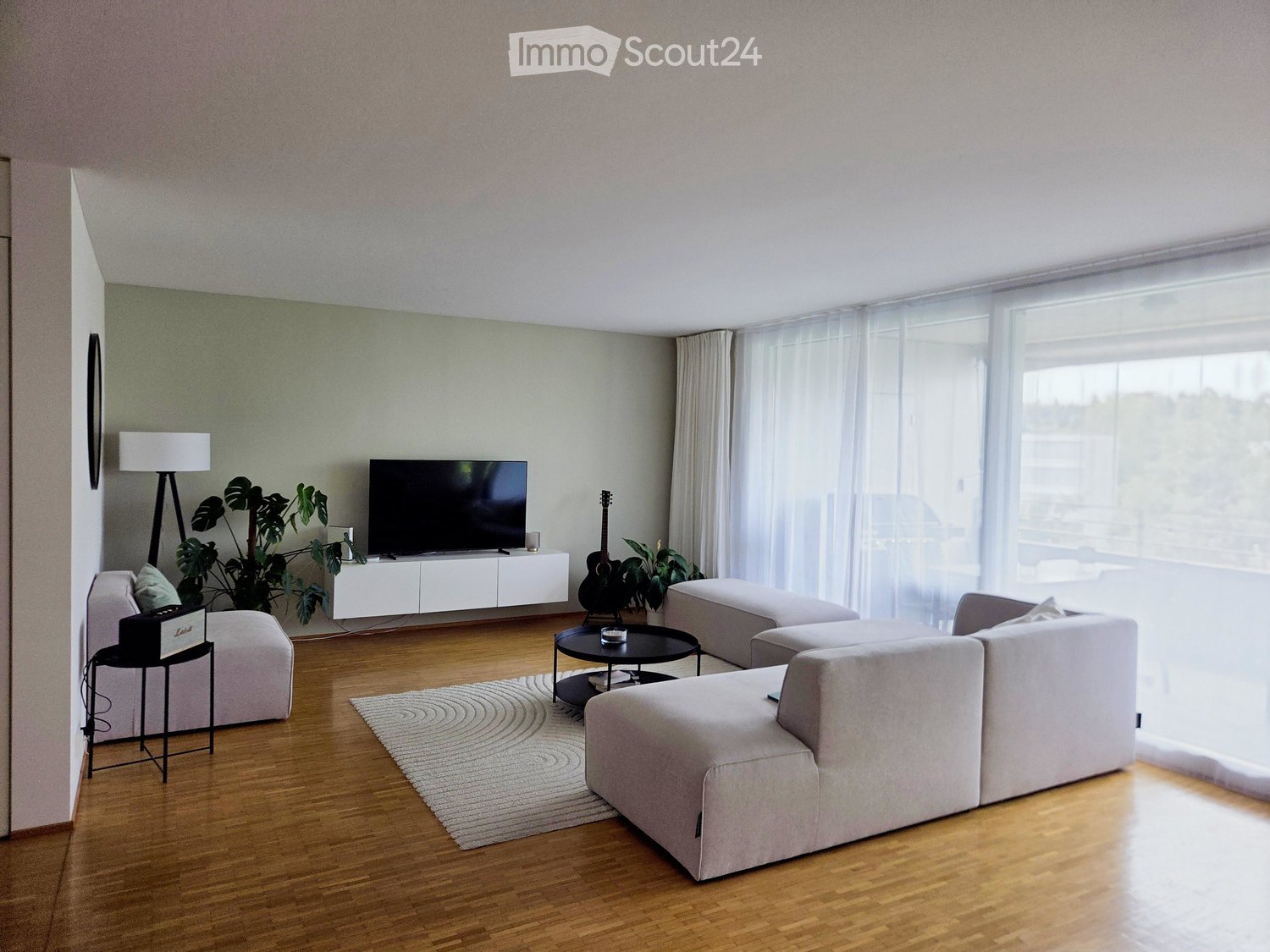Modern living room with light-colored furniture, a large sectional sofa, ottoman, coffee table, TV, and plants.