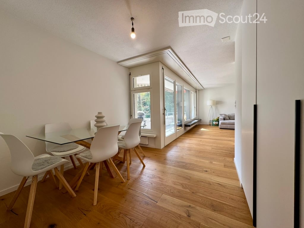 Bright, open-plan dining and living area with wooden flooring, large glass windows, a modern dining table with chairs, and a sofa in the background.