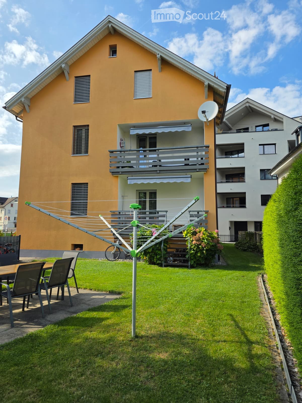 A well-maintained garden area in front of a two-story house with a balcony, featuring a clothesline, outdoor furniture, and a bicycle.