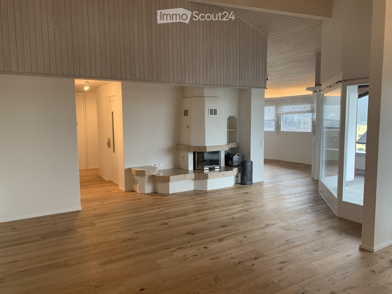 Empty room with wooden flooring, white walls, and a fireplace with a stone surround. The room has large windows and a glass door leading to another space.
