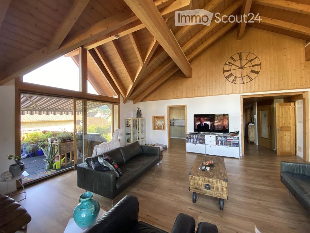Bright living room with wooden beams, large windows, and a view of the garden. Features a dark green sofa, coffee table, TV, and decorative items.