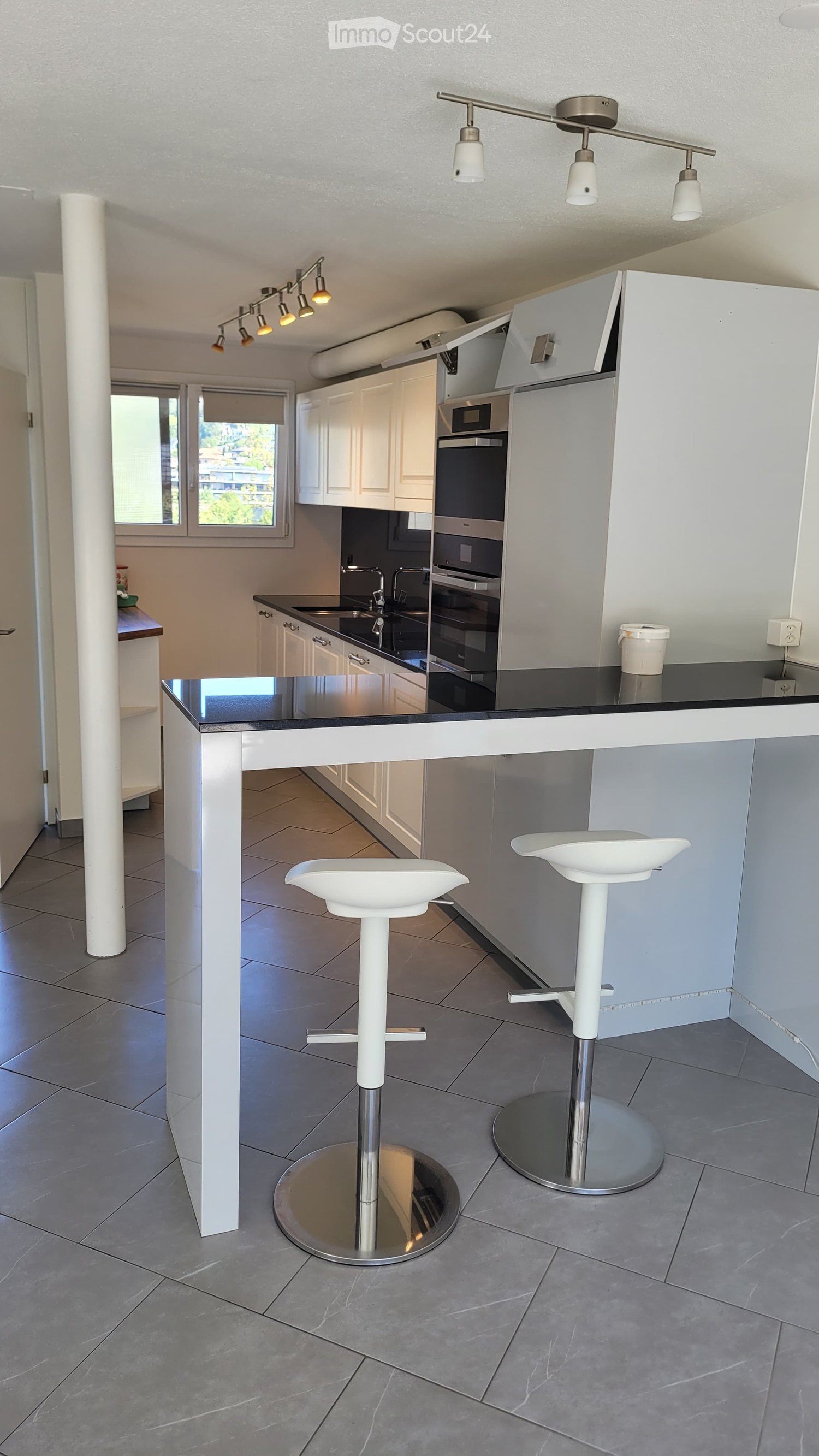 Modern kitchen with white cabinets, stainless steel appliances, black countertops, and two white bar stools at the island.