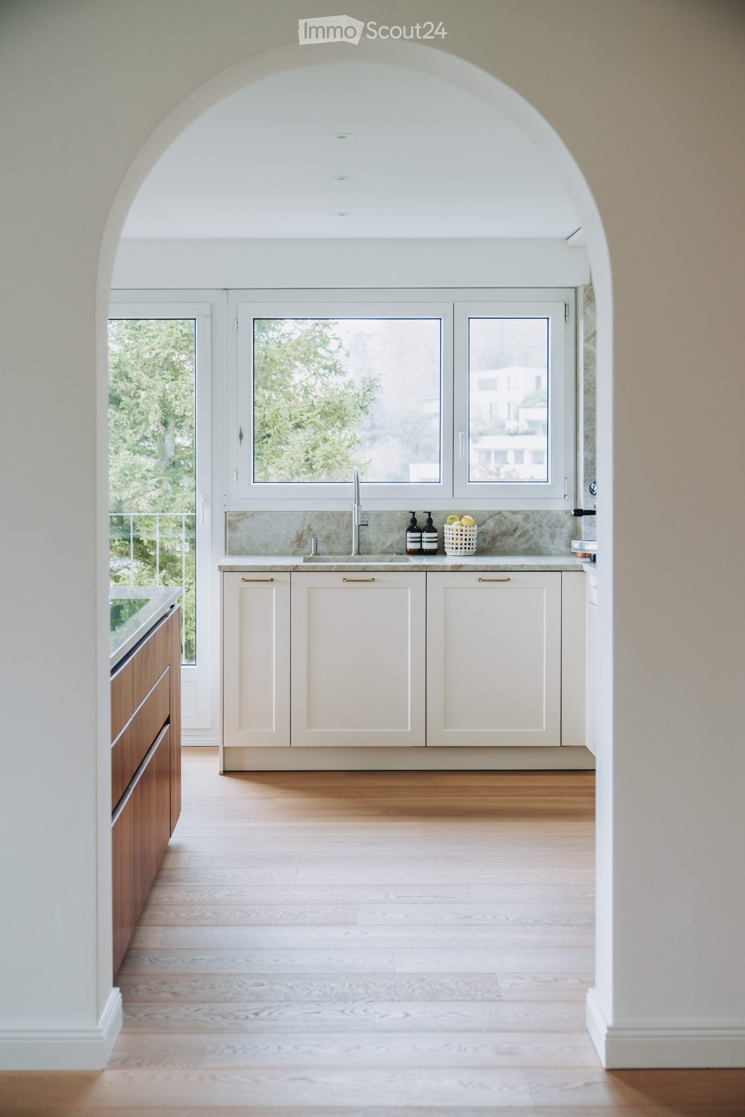 Modern kitchen with white cabinets, wooden flooring, and a large window overlooking greenery. Features a sink, faucet, and small decorative items on the counter.