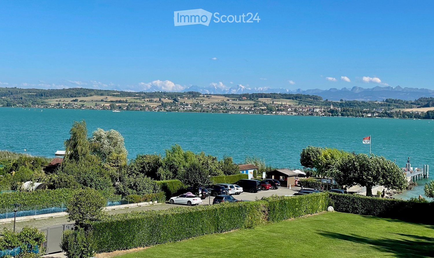 View of a lakeside area with a parking lot, small structures, and a flagpole, with a distant town and mountains in the background.