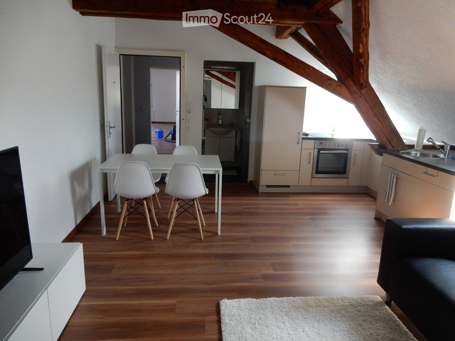Bright, open living space with wooden flooring, white dining table and chairs, modern kitchen area with cabinets and appliances, and a black leather sofa.
