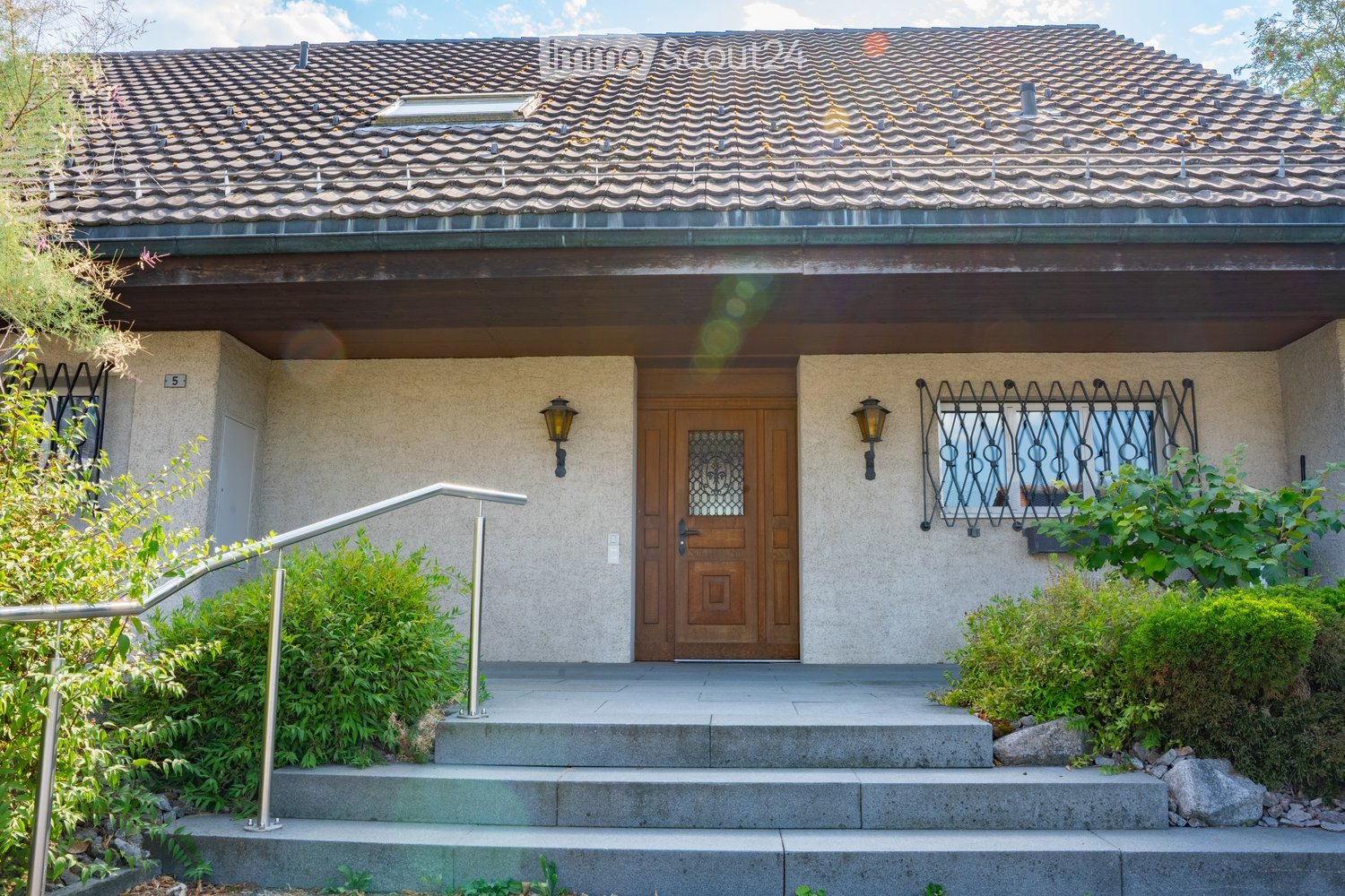 Two-story house with a tiled roof, beige facade, wooden front door, decorative window grilles, and small garden area with steps leading to the entrance.