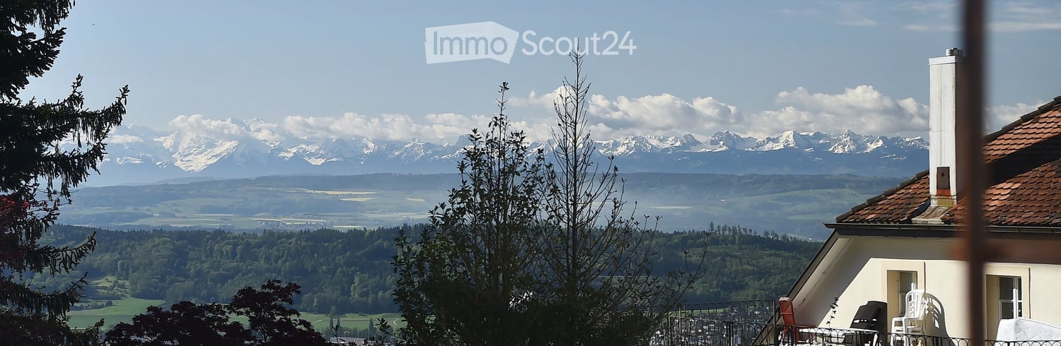 View from a balcony or terrace showing a house with a red-tiled roof, surrounded by trees, with a panoramic mountain range in the background.