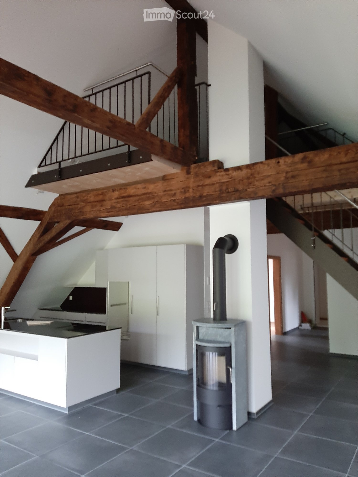 Modern kitchen area with white cabinets, a kitchen island, and a wood stove, featuring exposed wooden beams and a staircase leading to an upper level.