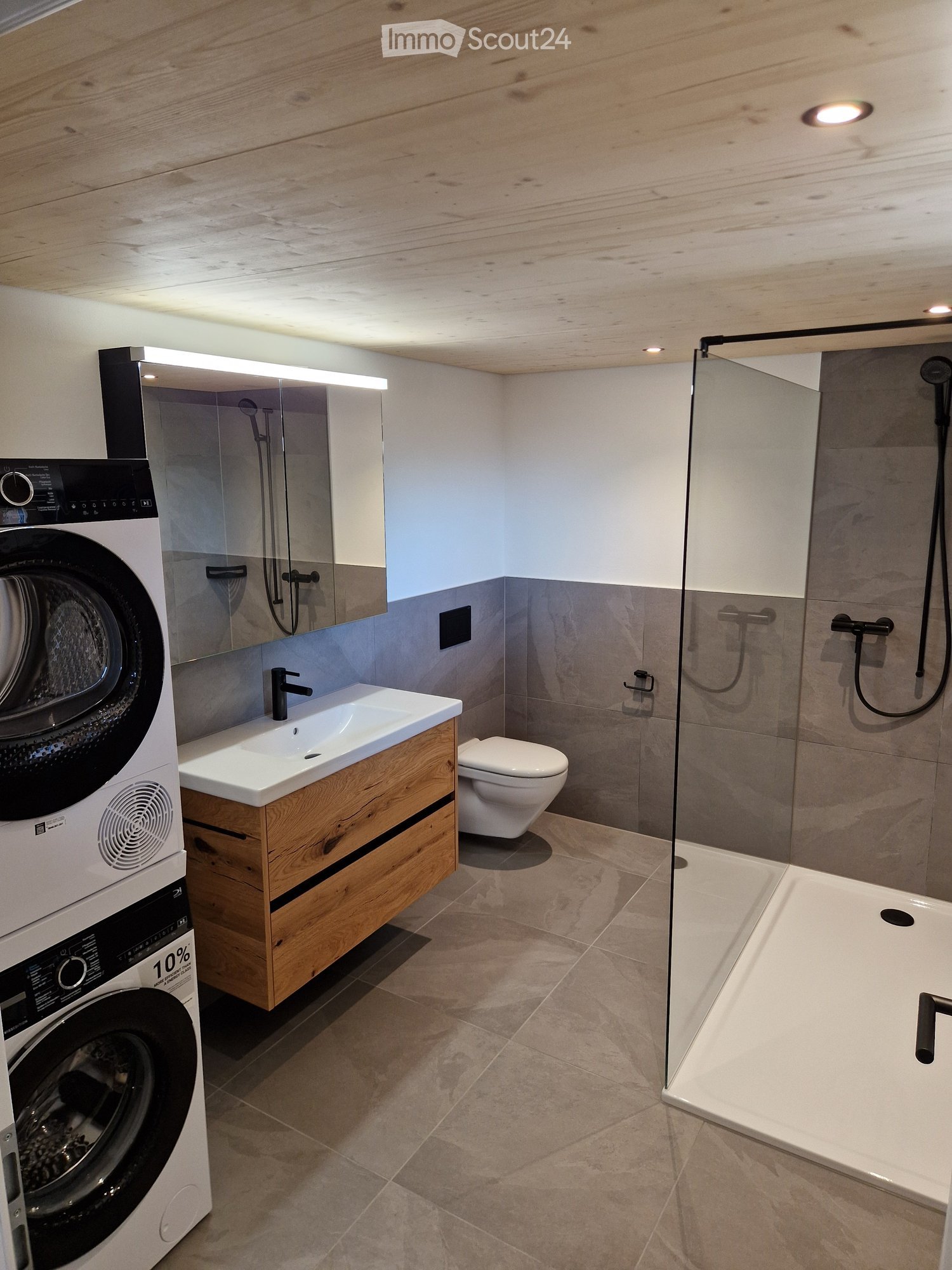 Modern bathroom with a wooden vanity, sink, toilet, bathtub, and washing machine. Features include a glass shower enclosure, wooden ceiling, and tiled floor.