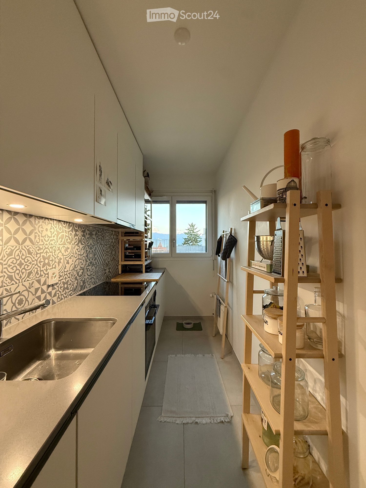 Modern kitchen with white cabinets, stainless steel sink, wooden shelf with jars and kitchen items, and a view of the balcony.