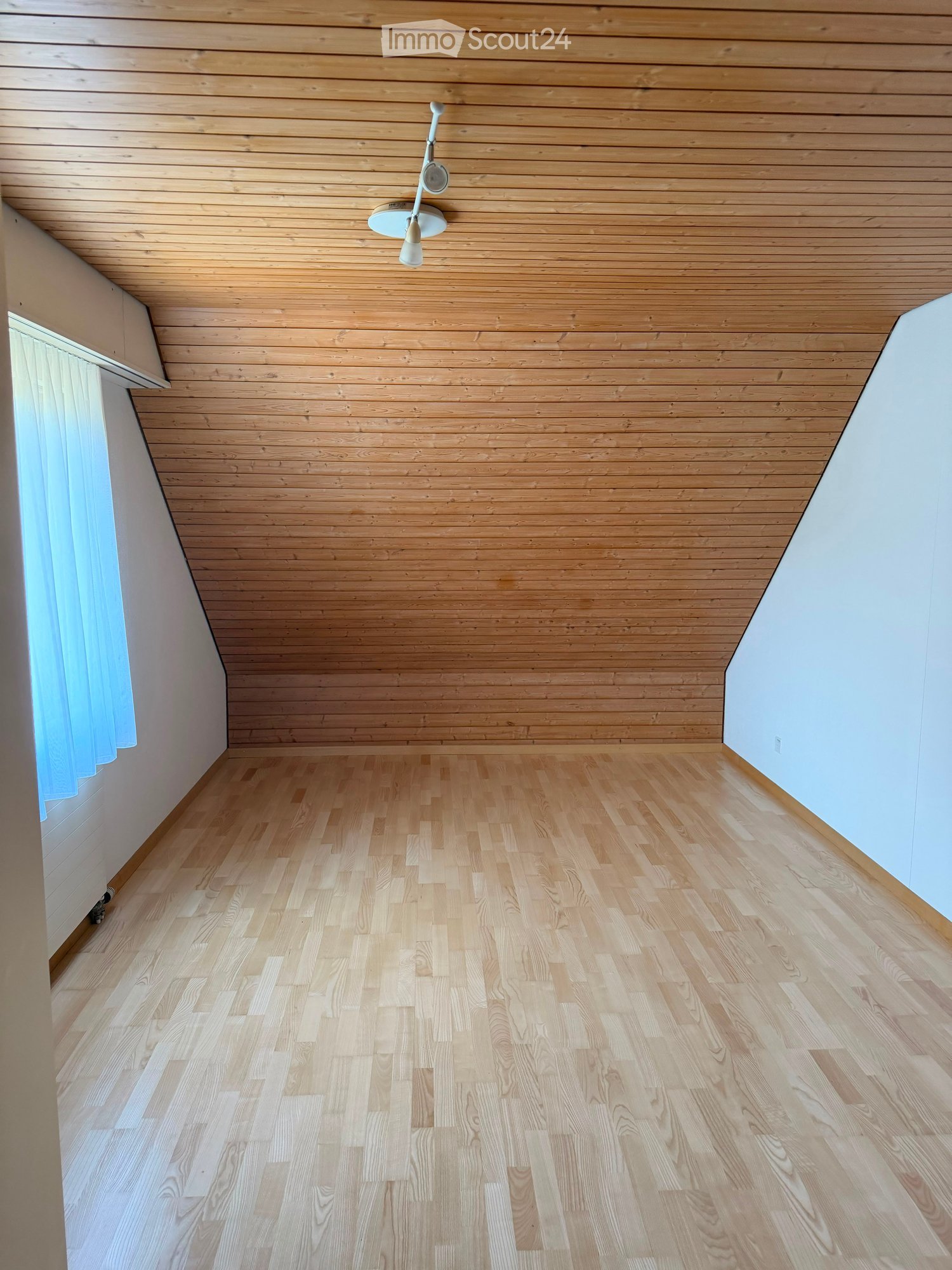 Empty room with light wooden flooring and paneled wooden ceiling, white walls, and a small window with a curtain.