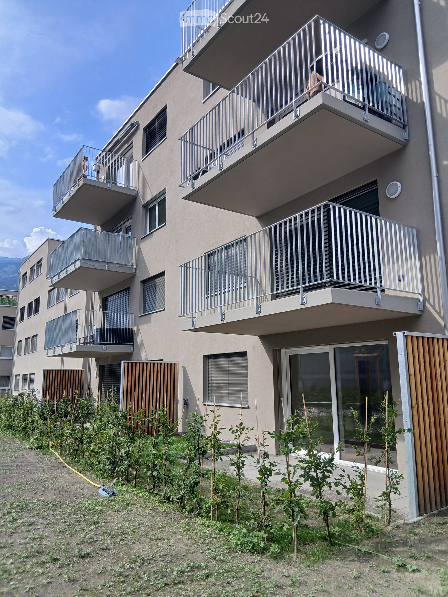 Modern multi-story residential building with multiple balconies, large windows, and a small garden area with young plants in front.