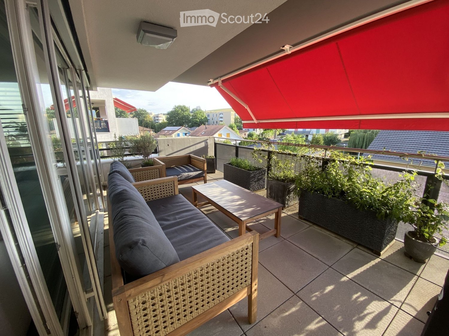 Balcony with a wooden sofa, a small table, a chair, and several potted plants. Features a red awning and a view of the surrounding area.