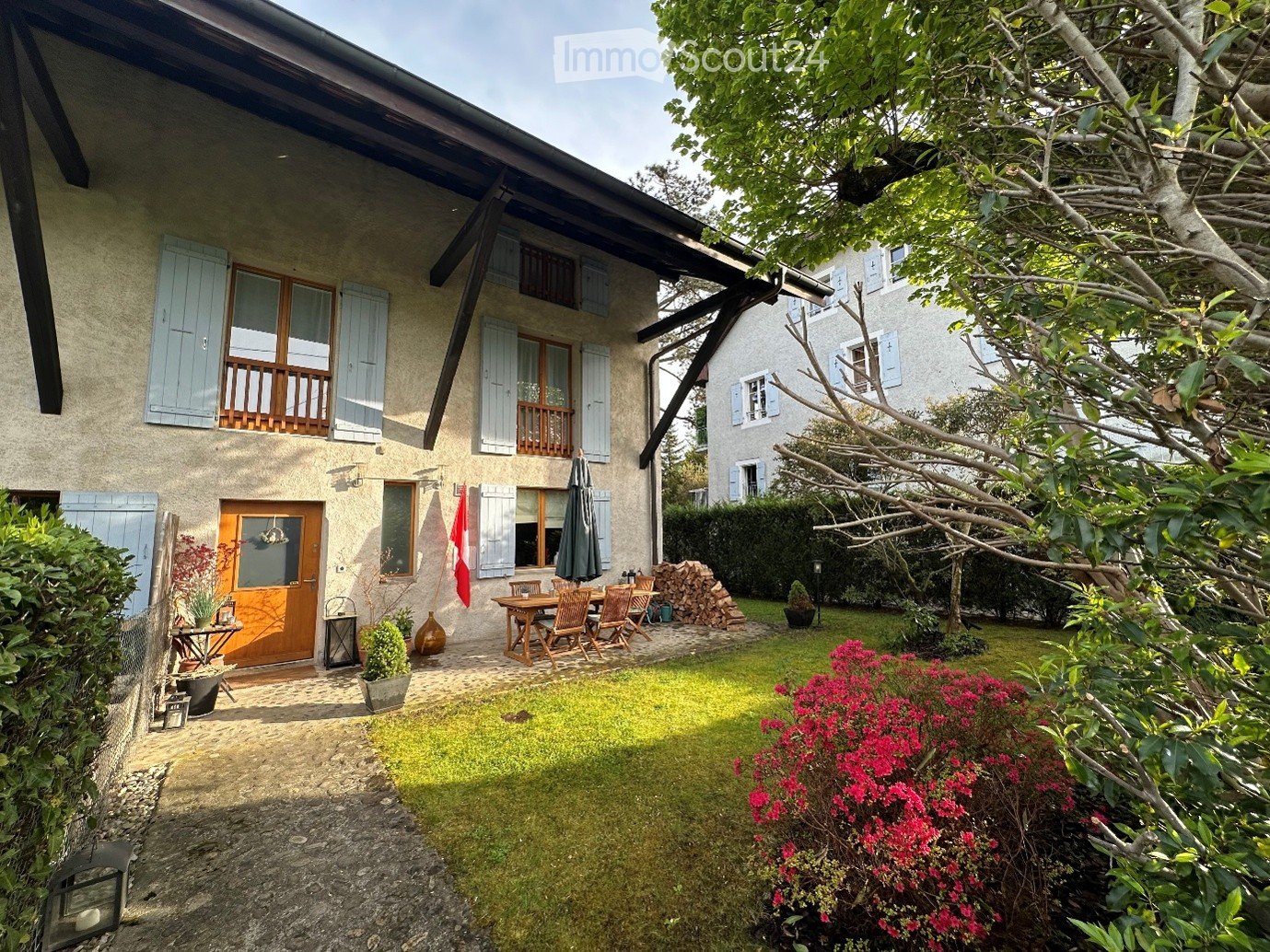 A well-maintained garden area with a wooden dining set, a closed umbrella, a stack of firewood, potted plants, and a flag. The garden is adjacent to a two-story house with light blue shutters and a wooden balcony.