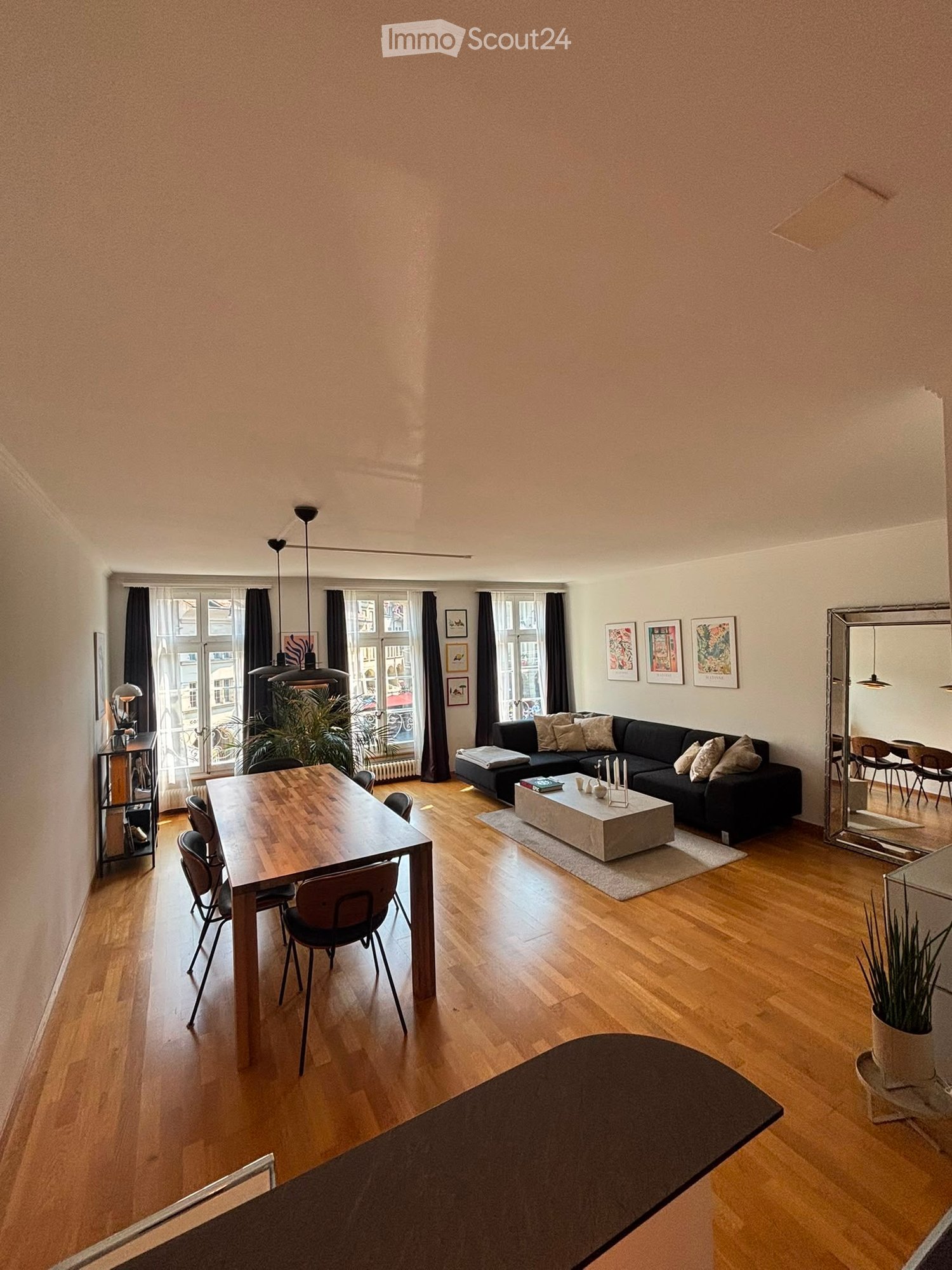 Open-plan living and dining area with wooden flooring, large windows, a black sectional sofa, a coffee table, a dining table with chairs, and a large mirror reflecting a dining area.