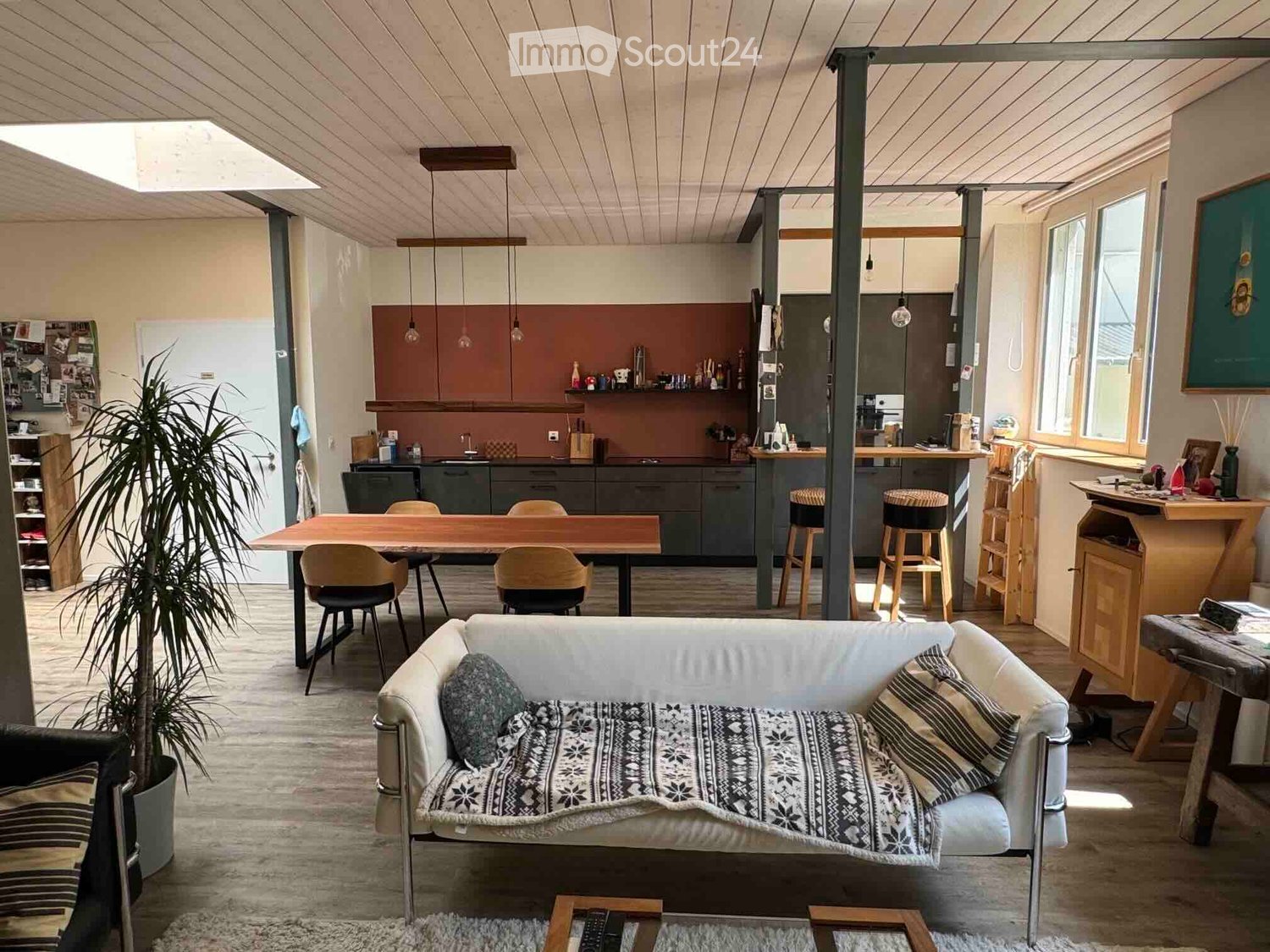Open-plan living and dining area with a sofa, dining table, chairs, kitchen area, pendant lights, and a plant.