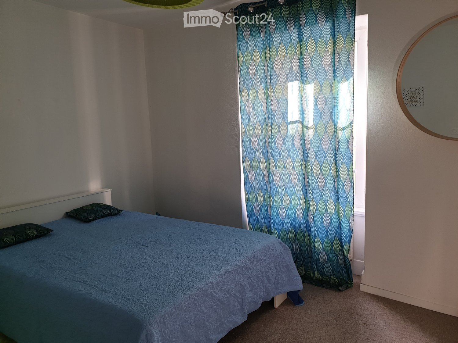 Bedroom with a blue bedspread, pillows, a mirror, and a window with patterned curtains.