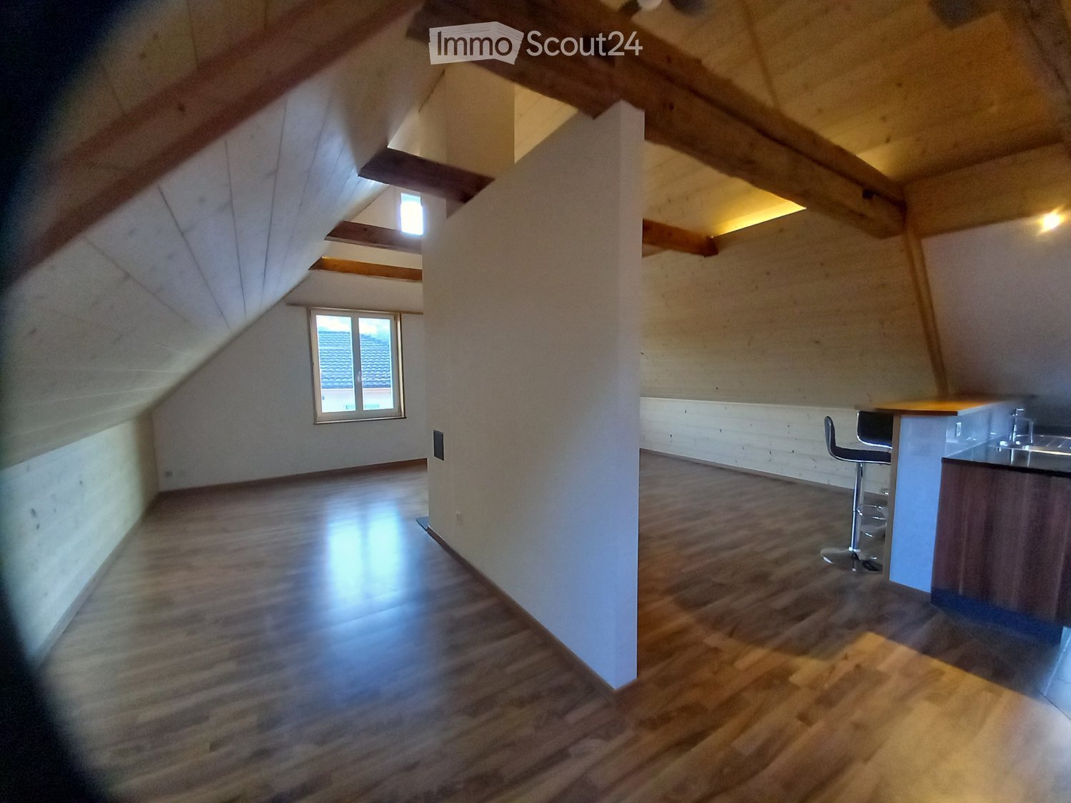 Bright, open attic space with wooden flooring, exposed wooden beams, and a small kitchen area featuring a counter and a stool.