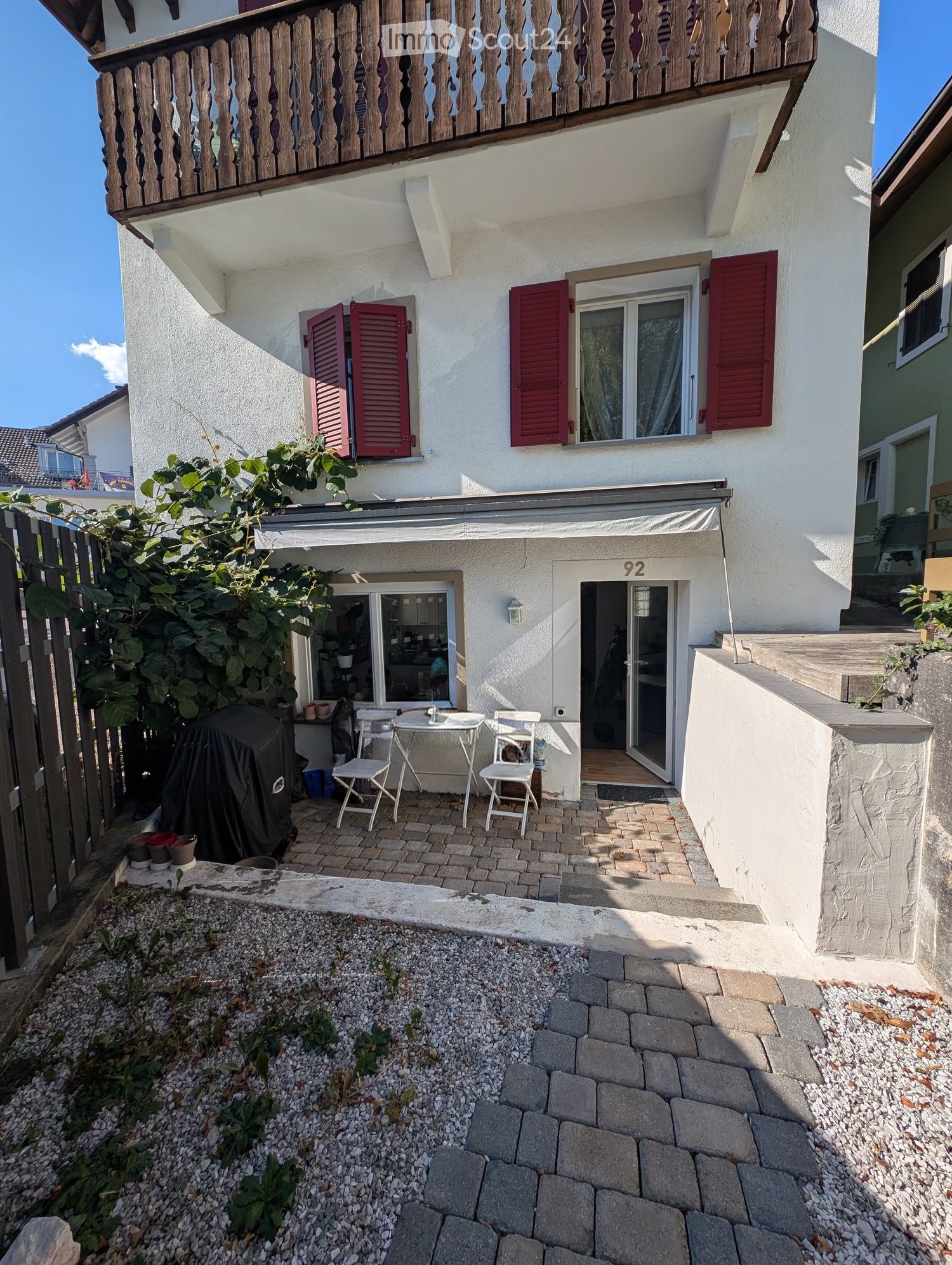 Small outdoor patio area with a table, chairs, and a covered grill, adjacent to a two-story house with red shutters and a balcony.