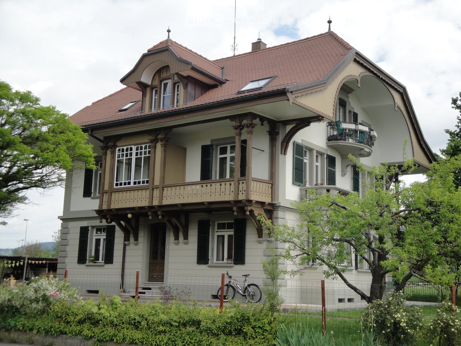 Two-story house with a red-tiled roof, white and beige exterior, multiple windows, balconies, and a bicycle parked near the entrance.