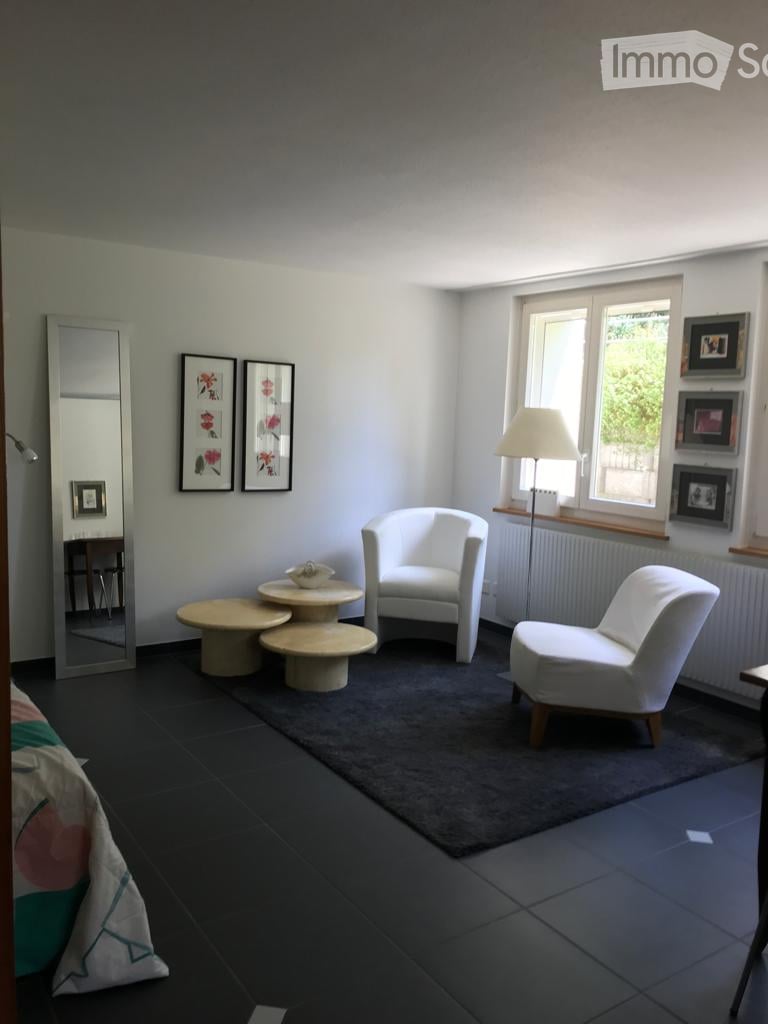 A modern, minimalist living space with white walls, dark flooring, and large windows. Features include a white armchair, small round tables, a floor lamp, and decorative picture frames.