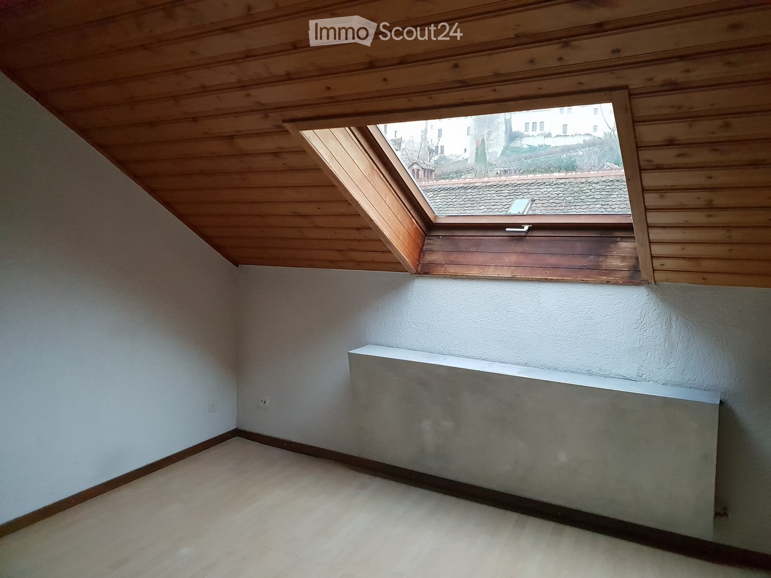 Empty attic room with wooden slatted ceiling, skylight window, white walls, and a radiator.