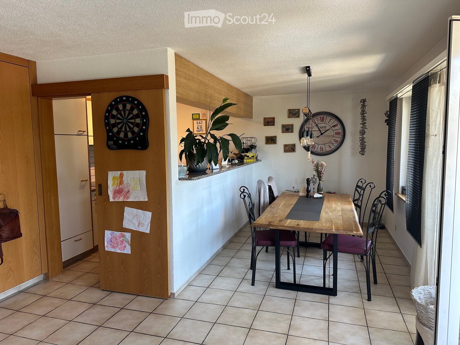 A modern dining area with a wooden table, four chairs, a large clock on the wall, a dartboard, and a potted plant on the counter.