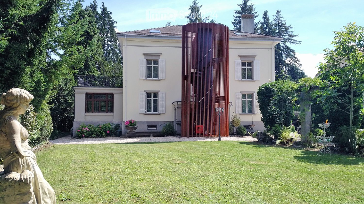 A well-maintained garden with a lawn, a statue, and a large house with white shutters and a red gate in the background.
