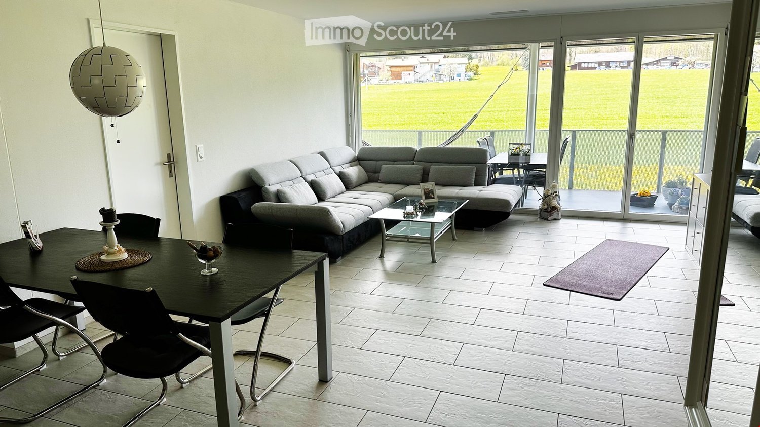 Modern living and dining area with a grey sectional sofa, glass coffee table, black dining table with chairs, and a hanging spherical lamp. Large sliding glass doors open to a view of a green field.