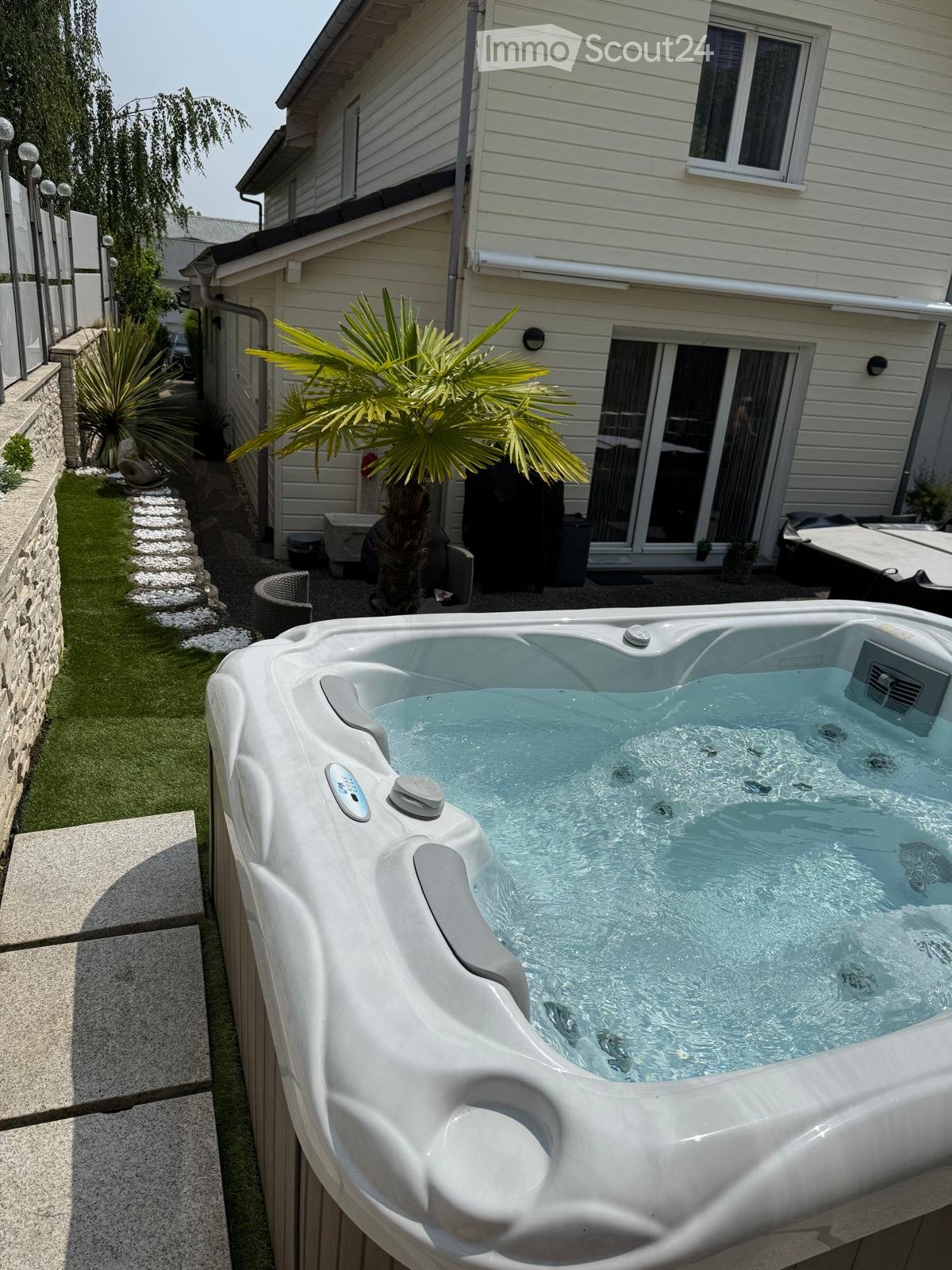Private garden area with a modern hot tub, surrounded by a stone wall, greenery, and a house with large windows and a sliding door.