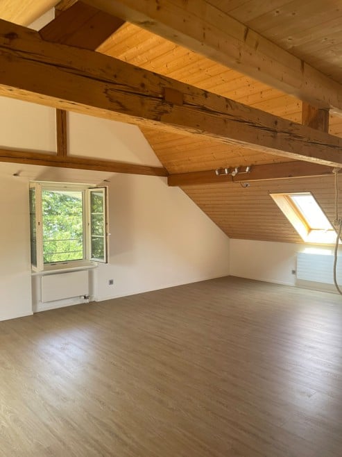 Empty attic room with wooden floor, exposed wooden beams, large windows, and skylight.