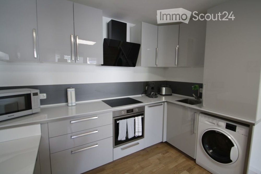 Modern kitchen with grey cabinets, white countertops, and stainless steel appliances including a microwave, coffee maker, toaster, oven, and washing machine. Features a range hood and a sink.