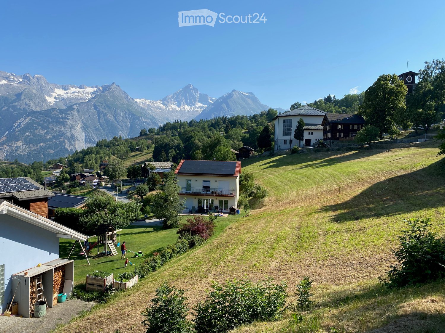A picturesque village scene with a house featuring a balcony, surrounded by green lawns, trees, and snow-capped mountains in the background.