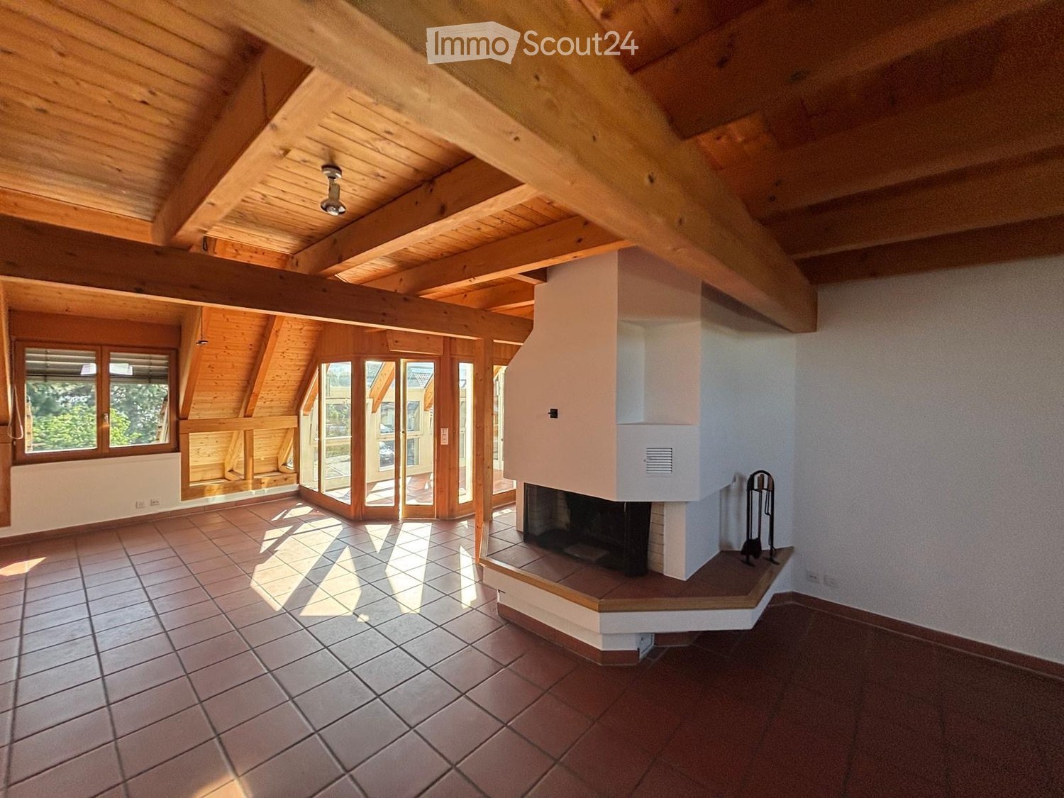 Bright, open room with exposed wooden beams, tiled flooring, large windows, and a fireplace. The space is empty, with no furniture or furnishings visible.