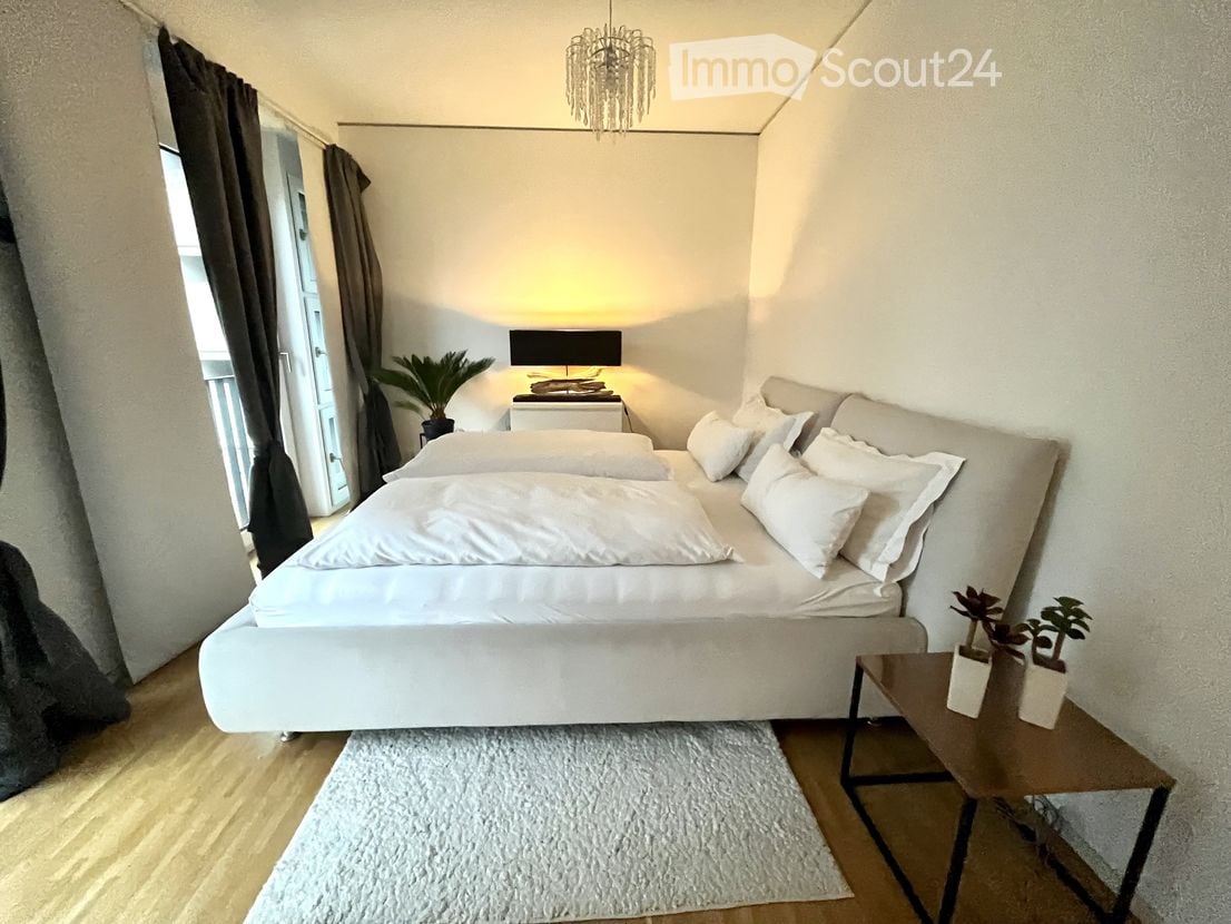 Modern bedroom with a large bed, nightstand, table lamp, side table with potted plants, and a view of a balcony.
