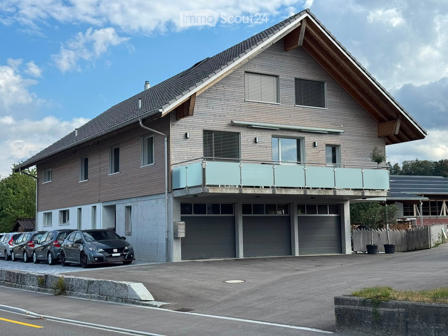 Two-story modern house with wooden siding, large windows, a balcony, and a garage with multiple parked cars.