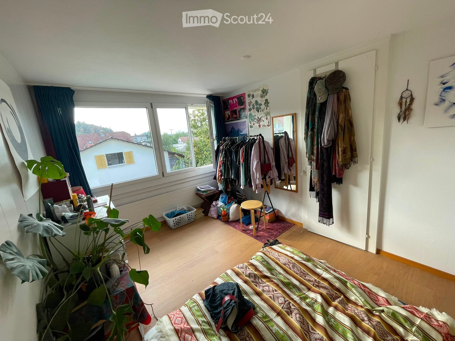 Cozy bedroom with wooden flooring, large windows offering a view, a bed with a colorful striped blanket, a small wooden stool, a clothing rack, a mirror, and a dreamcatcher on the wall.