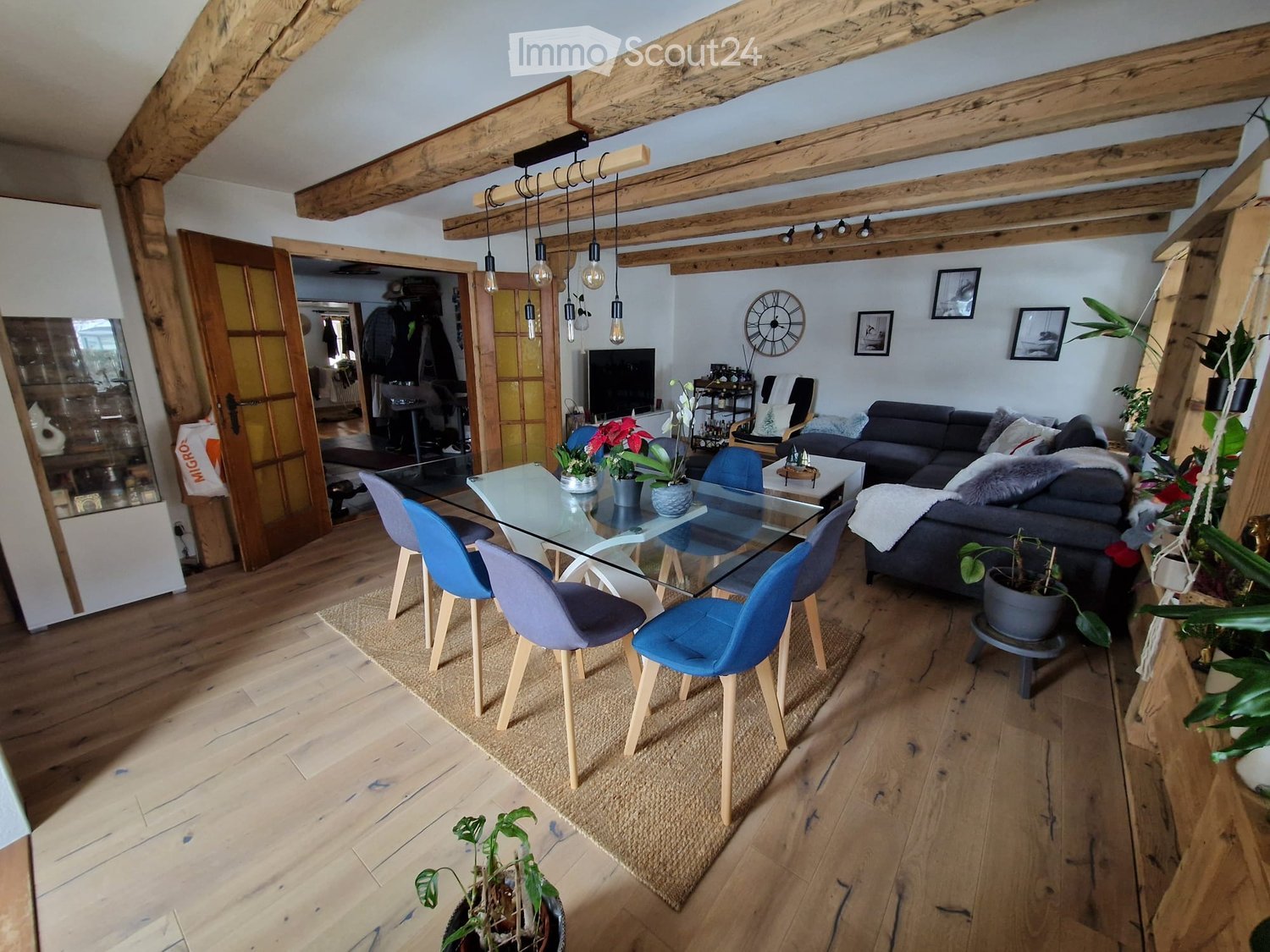 Cozy living and dining area with exposed wooden beams, hardwood flooring, a grey sectional sofa, a glass dining table with four blue chairs, and several potted plants.