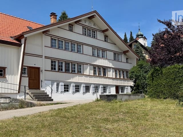 Two-story house with white siding, brown trim, and a red-tiled roof. Features multiple windows, a central entrance with steps, and is surrounded by a grassy area.