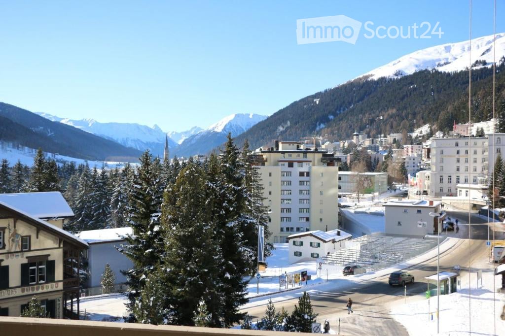 Panoramic view of a snowy town with mountains in the background, featuring buildings, trees, and a road.