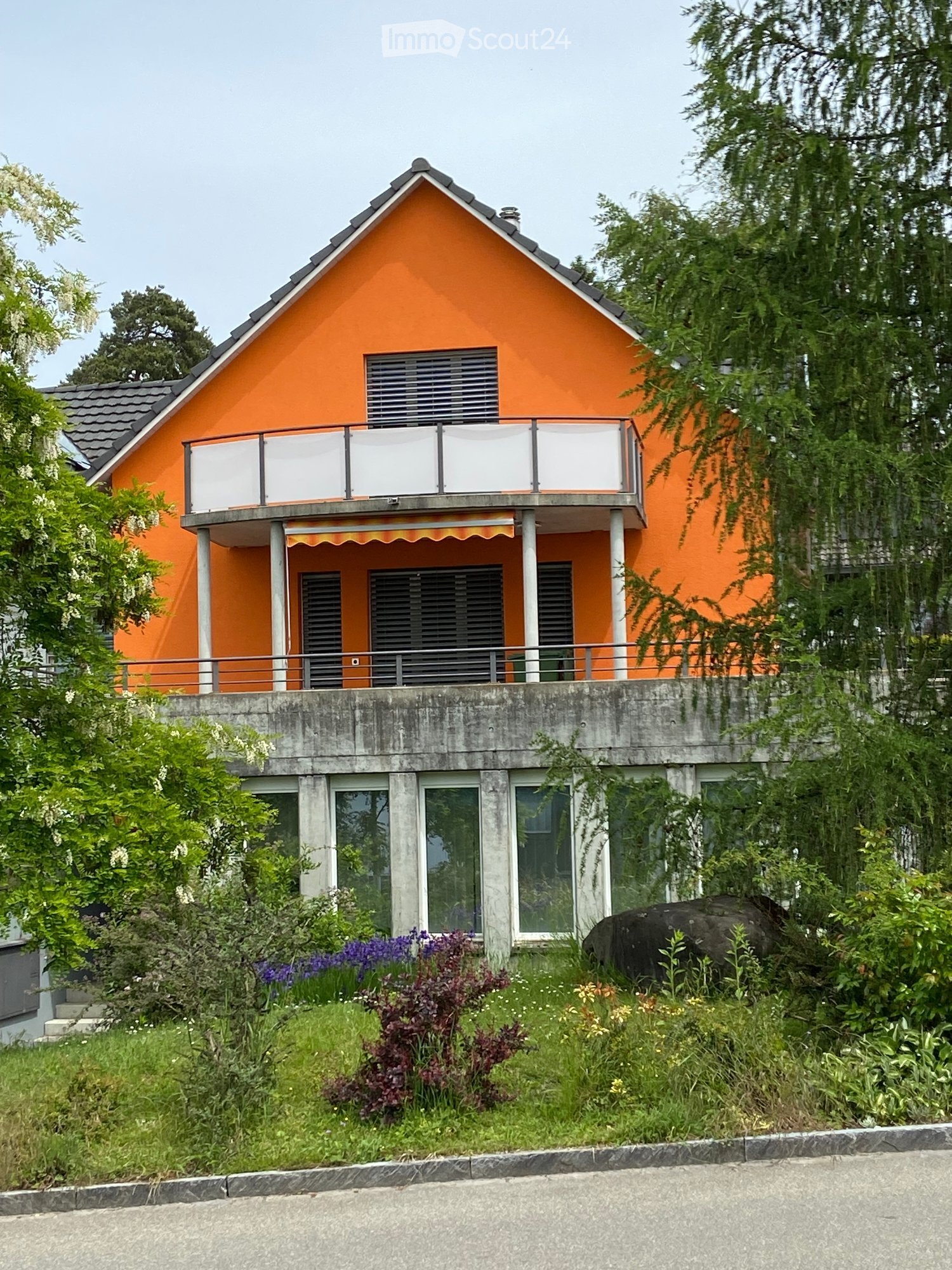 Two-story orange house with a raised ground floor, balcony, and surrounding greenery.