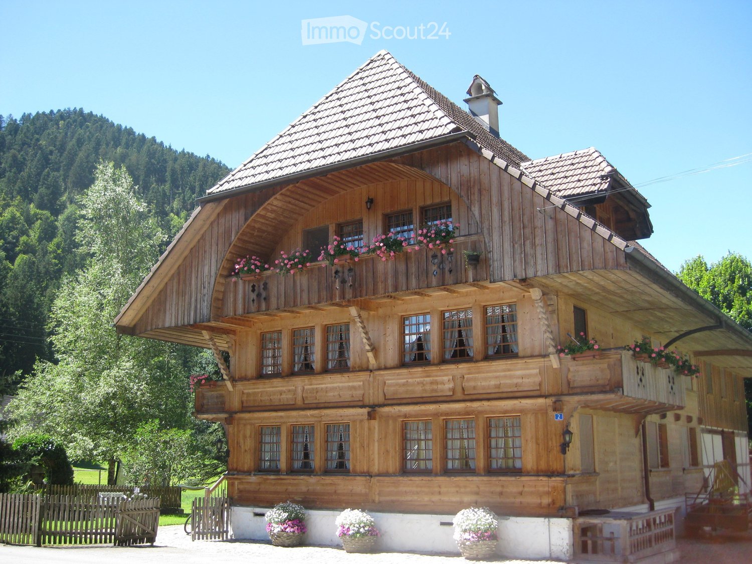 Traditional wooden house with a steep roof, multiple windows, and flower boxes on the balcony and ground level.