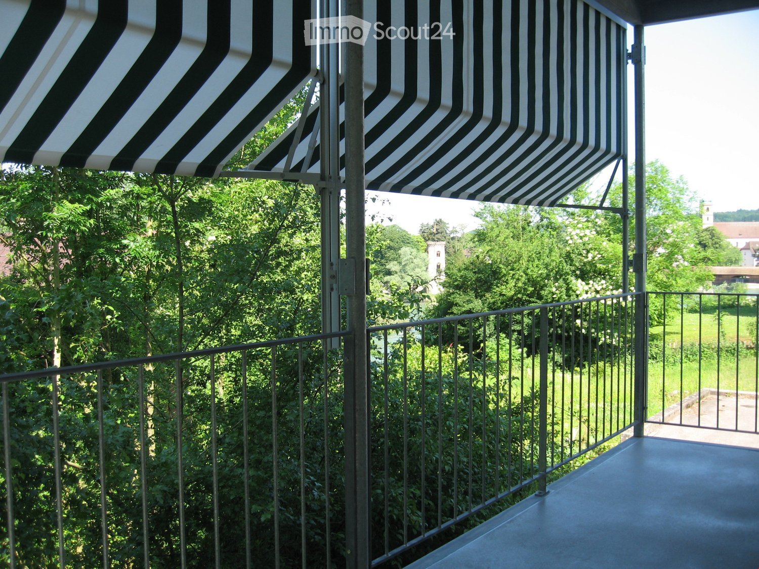 Balcony with metal railing, striped awning, and view of greenery and a distant church tower.