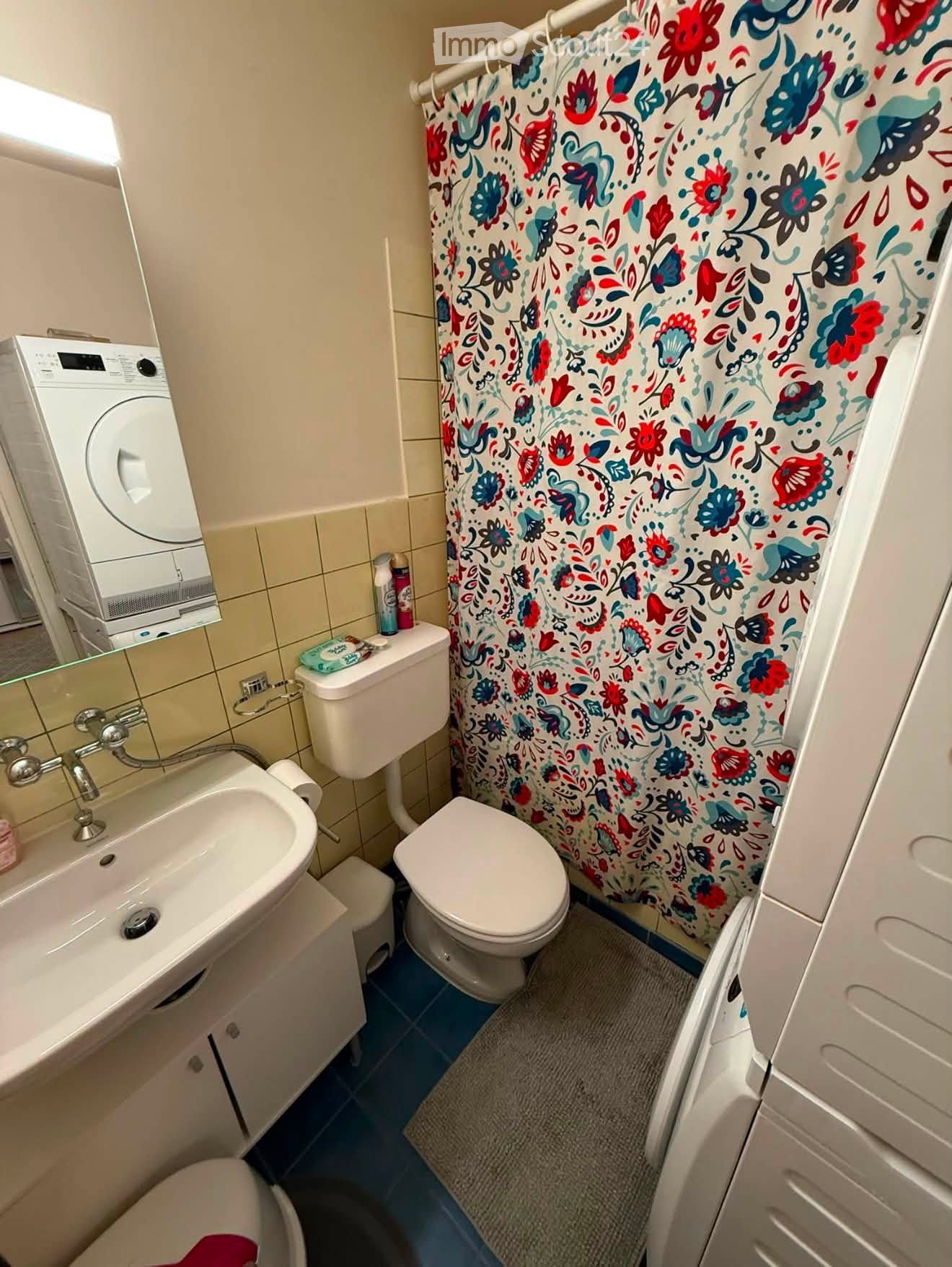 Compact bathroom with a sink, toilet, shower curtain, washing machine, and a small cabinet.