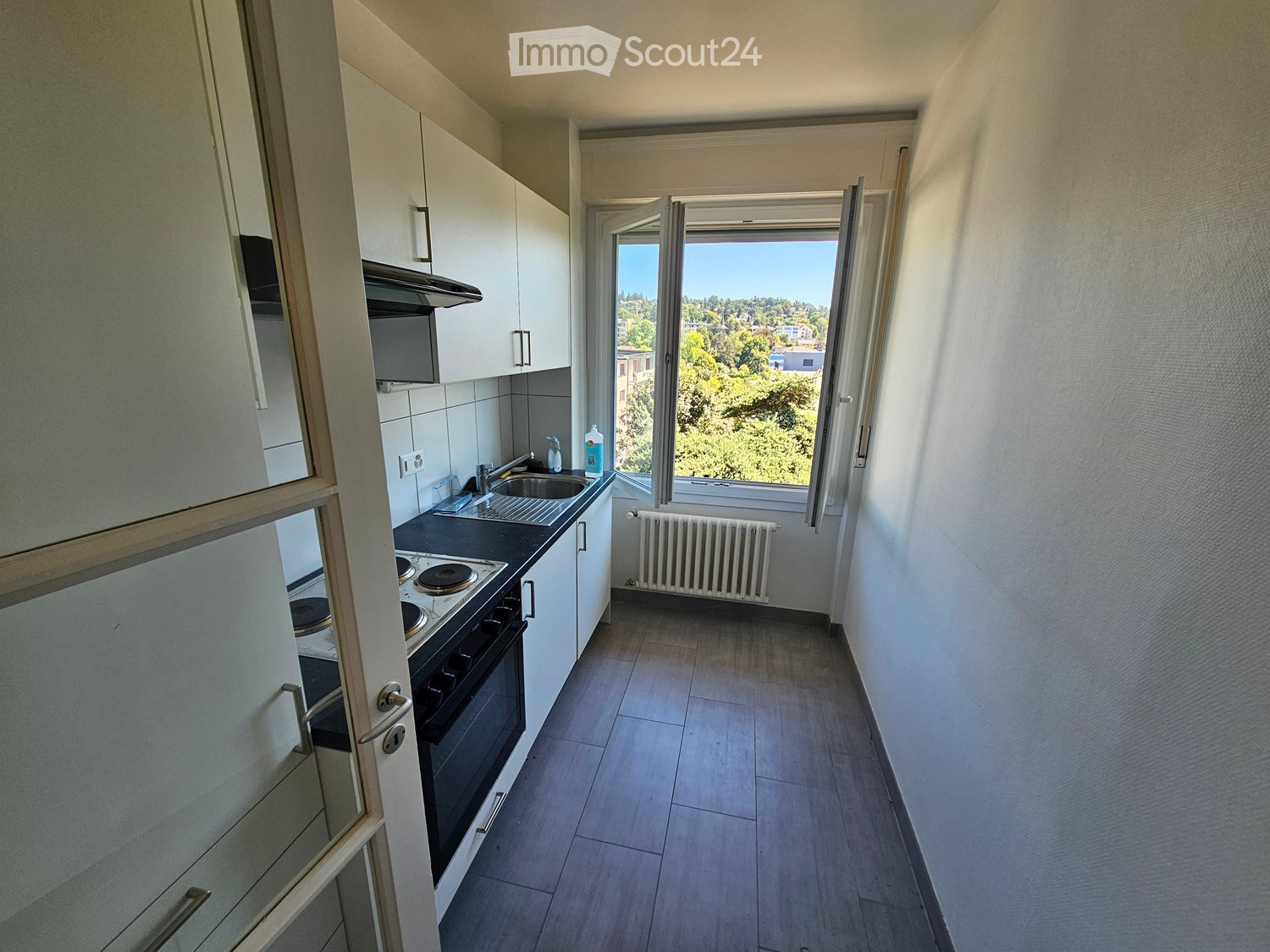 Modern kitchen with white cabinets, a black countertop, a stove, an oven, and a sink. The room has wooden flooring and a large window offering a view of greenery outside.