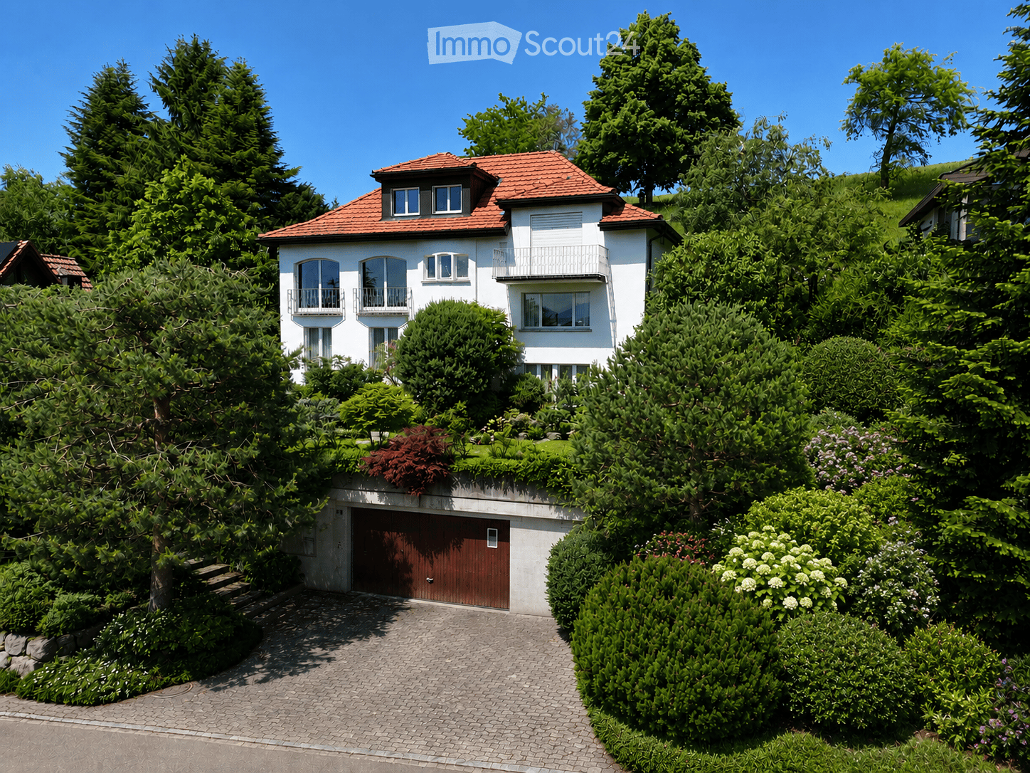 Two-story white house with red-tiled roof, multiple windows, balconies, and a garage surrounded by lush greenery and trees.